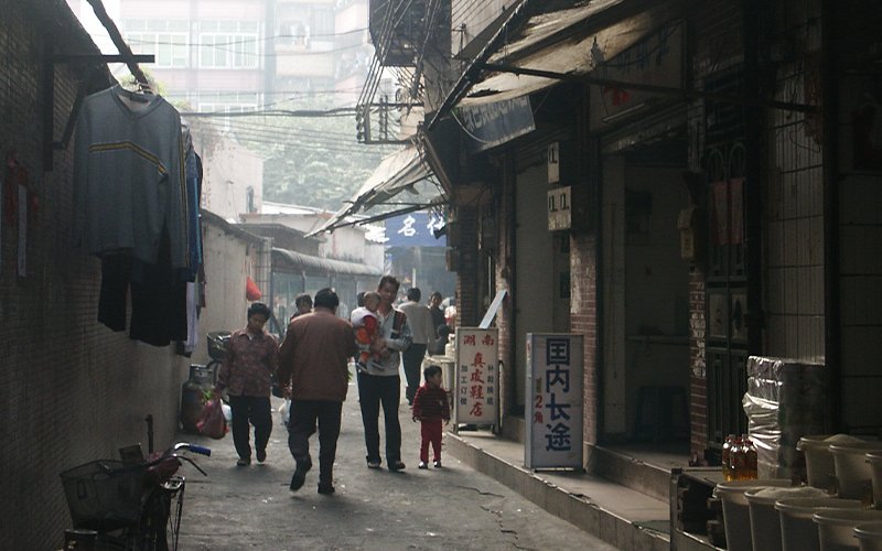 A family walks along a run down street in China, signs and clothes are displayed outside of storefronts on either side of the street.
