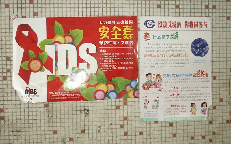 AIDS prevention signs hang on a faded and chipped white and peach tile wall.