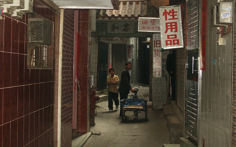 A man in dark clothes with a cart stands in a dark alley, surrounded by signs advertising goods and services. Another man walks past him on the left.