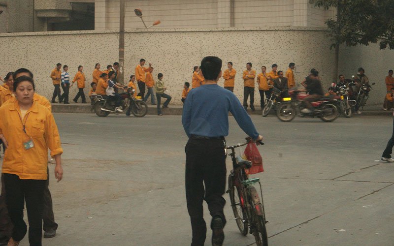 A man in a blue shirt and black slacks guides and bicycle toward a group of workers in bright yellow jackets outside of a building in China.