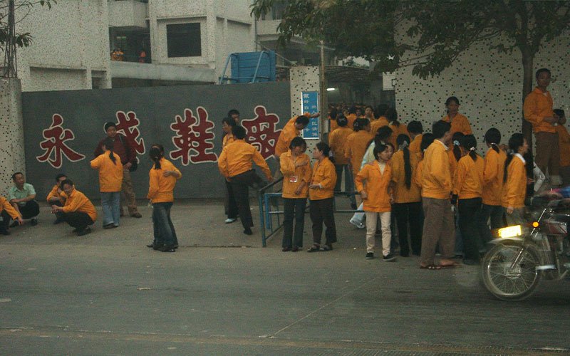 A group of workers in bright yellow jackets stand outside a building in China, in front of a wall with red Chinese lettering that translates to "Yongshan Shoes."