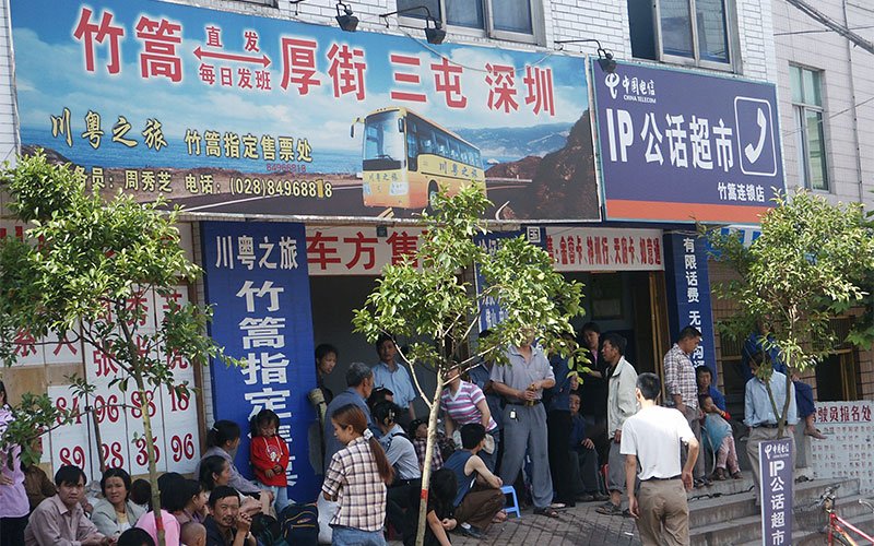 People stand on a street inside outside a storefront adorned with Chinese advertisements for a bus and transportation service.