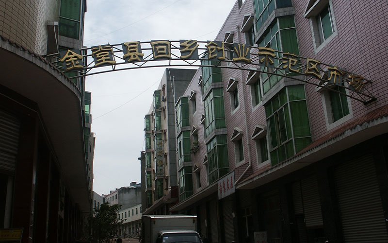 A street in China, lined with brick buildings with green windows. The street's entrance is decorated with a wrought iron arch overhead, displaying Chinese lettering.