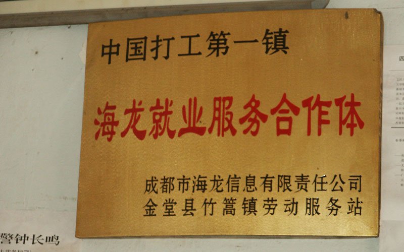 A gold sign with black and red Chinese lettering that translates to, "The first town for migrant workers in Chin/Hailong Employment Service Cooperation/Chengdu Hailong Information Co., Ltd. Jintang County Zhupeng Town Labor Service Station Four"