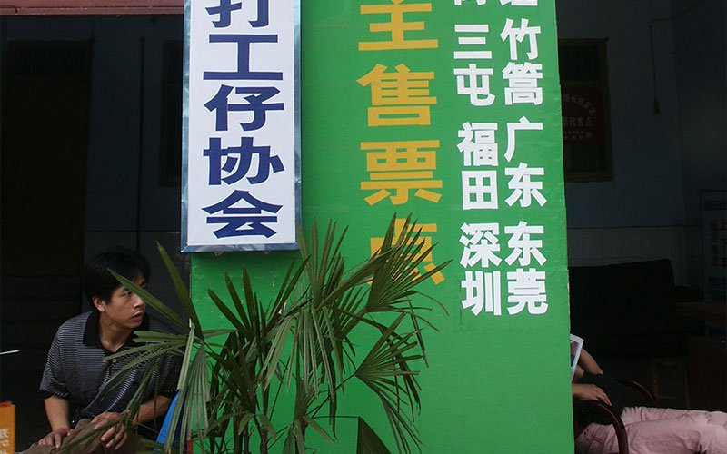 A man sits next to a bright green, white and yellow sign and palm plant in China. The sign, written in Chinese, promoted ticket sales and the Migrant Workers Population.