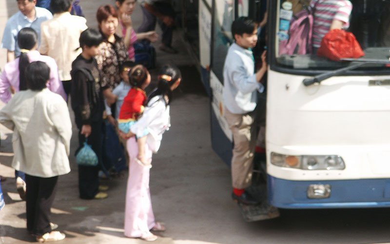 A crowd of people waits by a shuttlebus in China. The image is slight out-of-focus.