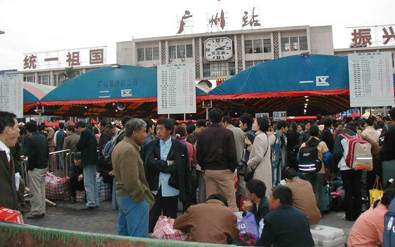 A crowd of people walk in front of Guangzhou Station in China. The station has three red and teal awnings out front, a clock on its facade and signs in Chinese that translate to, "Unify the motherland," "Guangzhou Station" and "Revitalize...," with the remaining text out of frame.