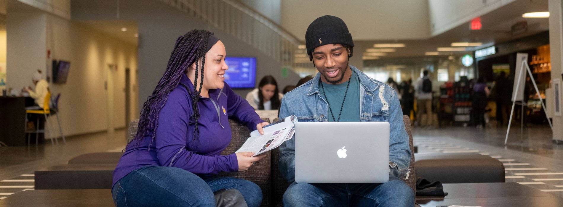 Two smiling students sit in Campus Center holding a laptop and some papers.