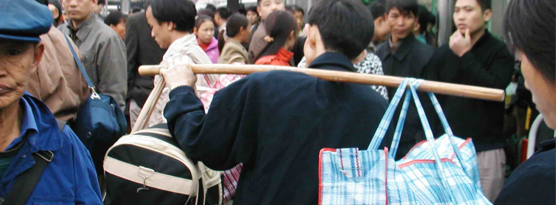 A man walks through a crowd in China, using a wooden pole to carry two large bags slung over his shoulder. One is a light-blue plaid pattern and the other is black with white trim.