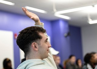 A student in a light-colored hoodie raises his hand in a crowded classroom.