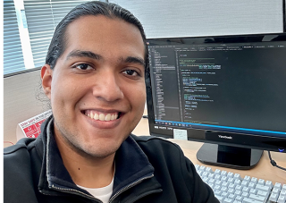 Jorge Gomez smiles in front of a computer monitor displaying programming text.