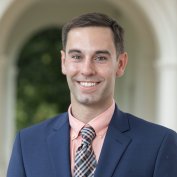 Dr. Jonathan Foster smiling blue suit jacket plaid tie peach shirt campus arch background.