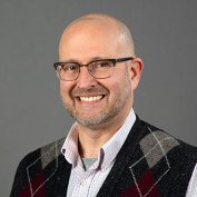 Justin Albohn smiles in glasses, a checked button up and argyle sweater vest against a gray backdrop