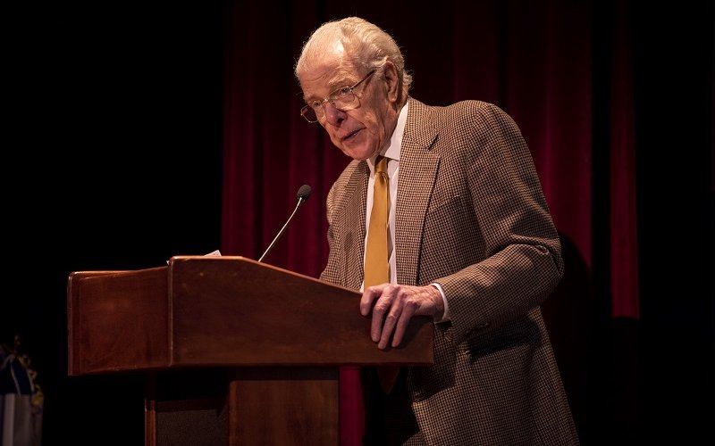 An older man in a tan suit coat and yellow tie leans into a dark wood lectern and speaks into a microphone