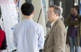 A student with his back to the camera describes his research poster to a man at a student research fair