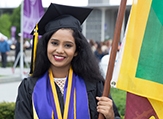 A female UAlbany degree recipient bears a flag and smiles at her May 2018 Commencement.