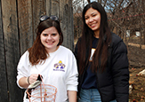 Two young women wearing UAlbany long-sleeve t-shirts smile after gathering eggs by a henhouse