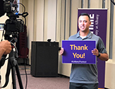 A student, being filmed, holds up a purple and gold Thank You sign