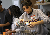 A young woman in a restaurant kitchen bends over a pot and spoons a white paste into it