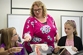 A young teacher stands, instructing two young girls sitting at a long table
