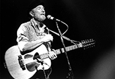 Pete Seeger at the microphone sings while holding guitar 