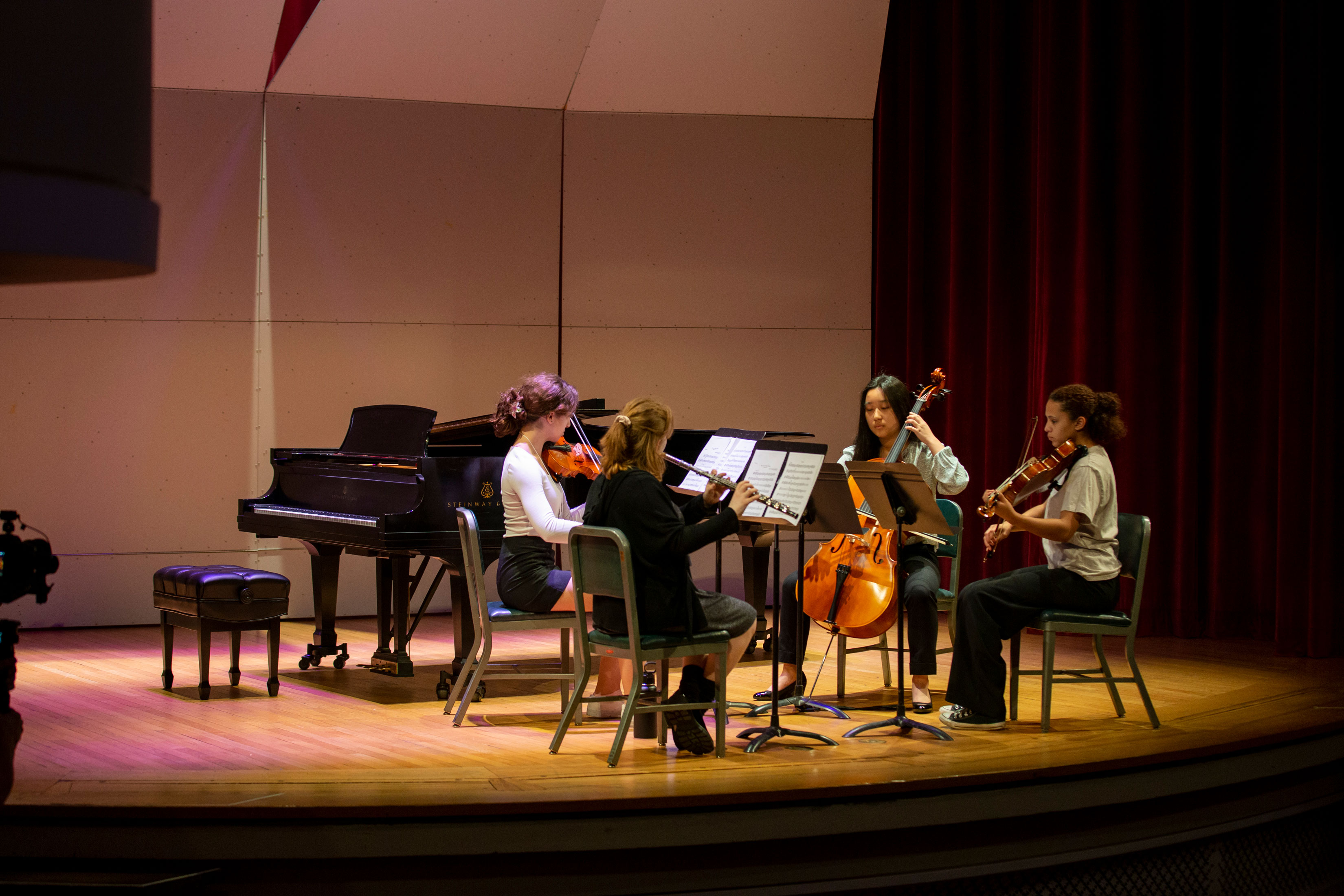 Four students, three with string instruments and one with a woodwind, sit on stage performing music.