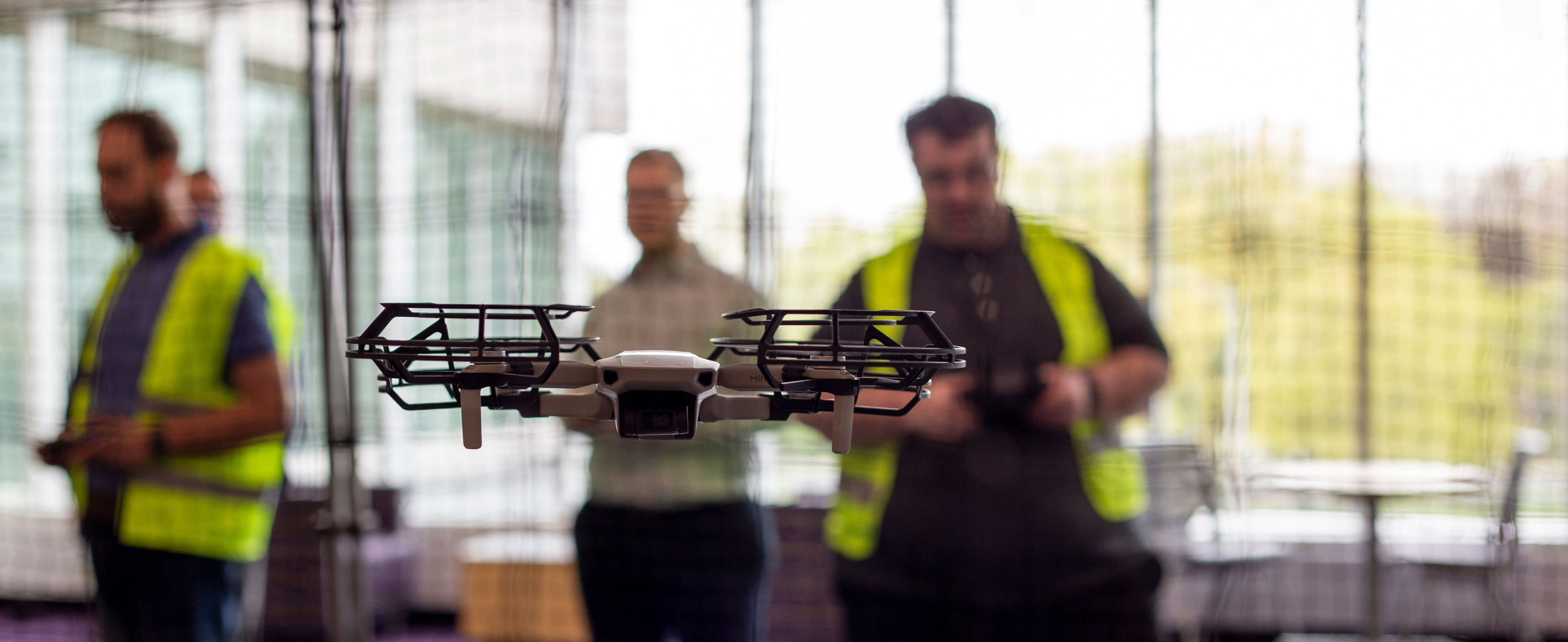 A drone hovers in the foreground while students wearing yellow vests stand behind netting and use a remote controller to guide the drone.