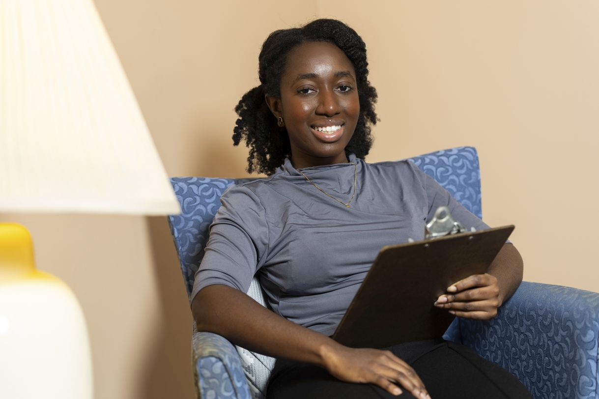 A young woman holding a clipboard smiles at the camera. She is seated in a blue upholstered chair in a warmly lit room. 