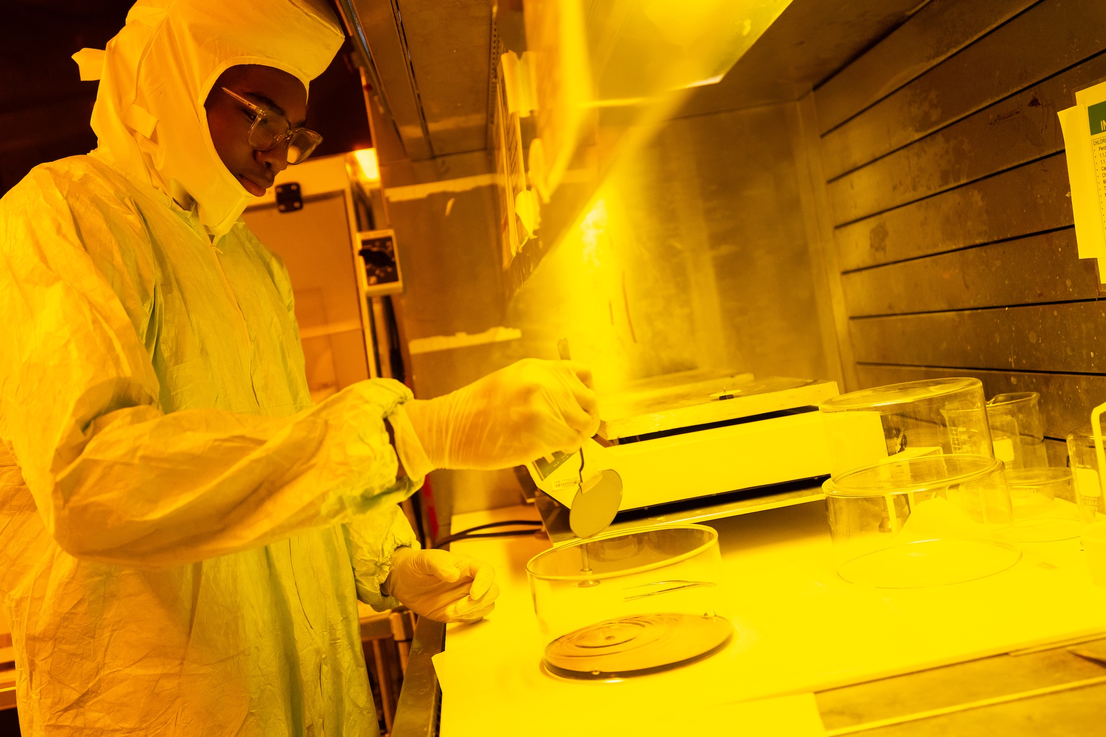 Student in protective bunny suit holds a semiconductor wafer with an instrument over a bowl. 