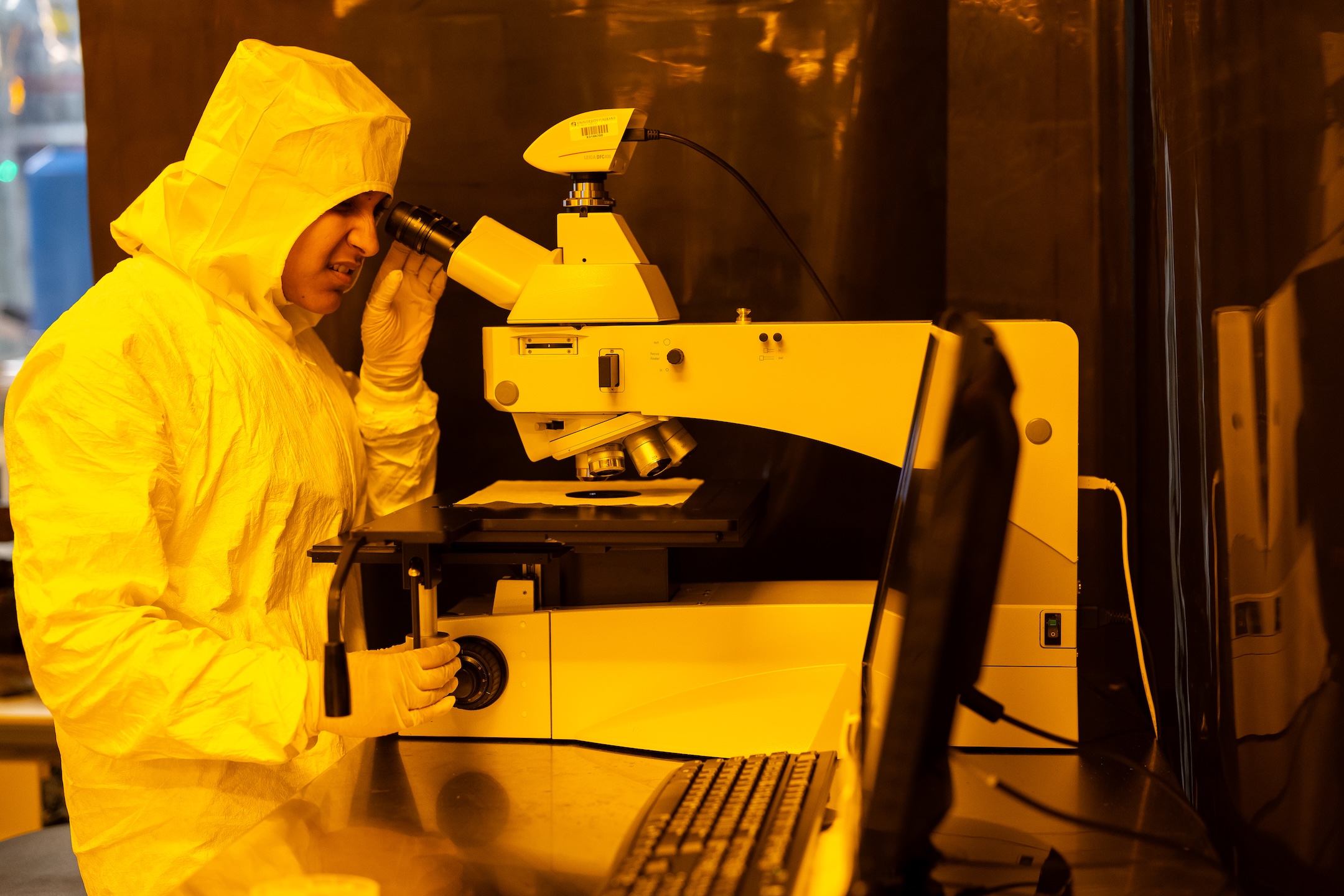 Student in protective bunny suit peers into a microscope inside lab.