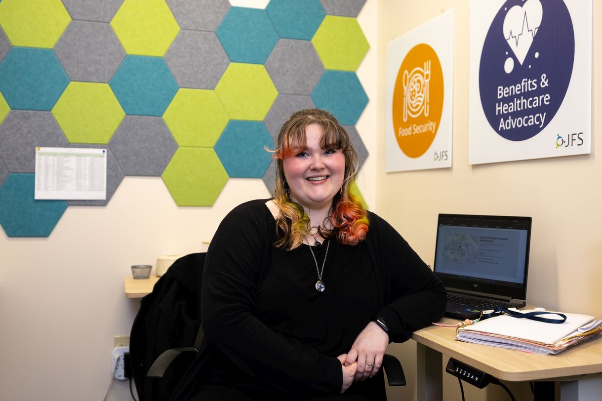 A young woman with rainbow hair smiles for a portrait while seated at a desk. Signs on the wall say "food security" and "benefits & advocacy".