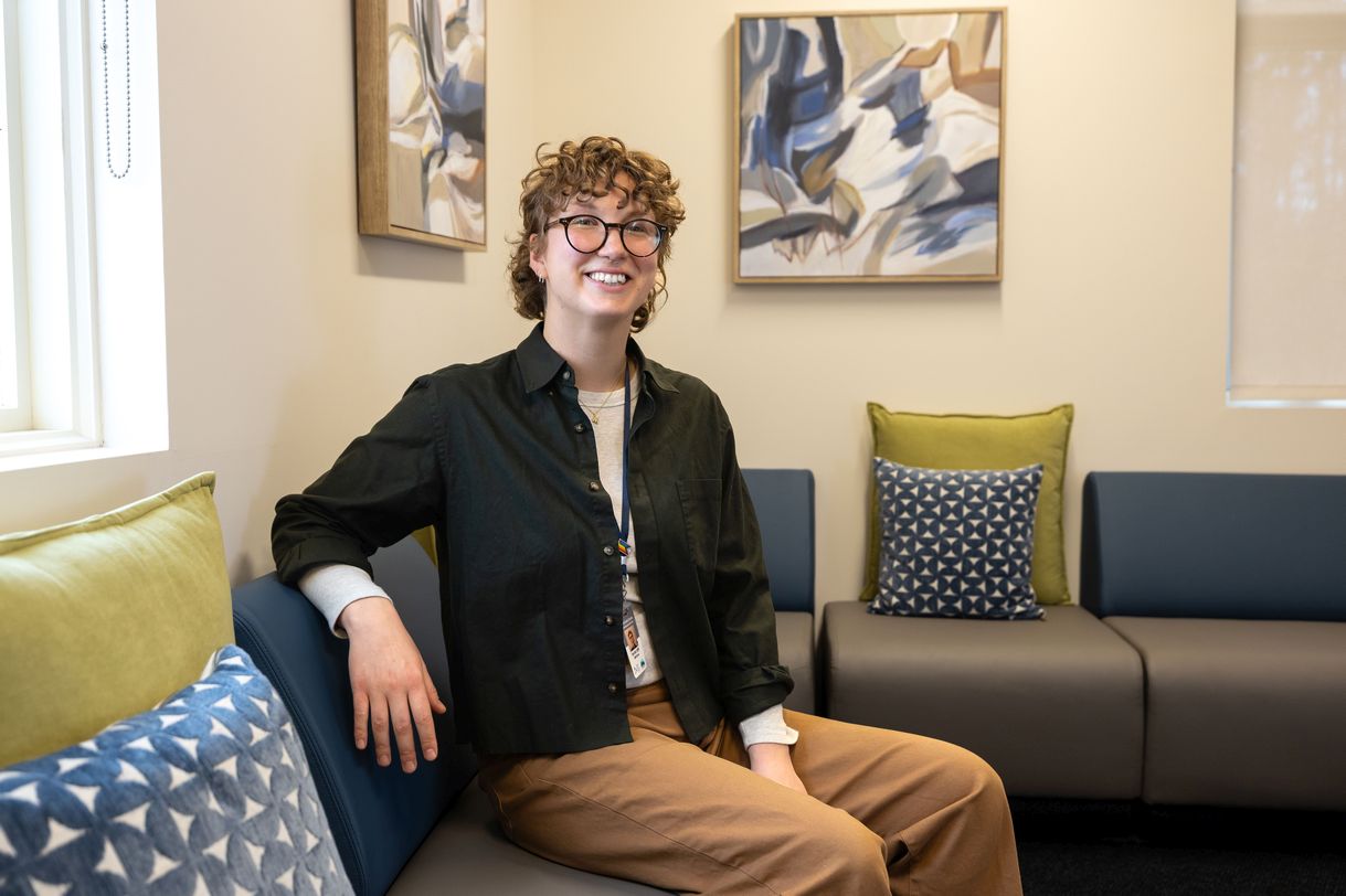 A young person wearing glasses and a green button down shirt sits on a sofa in a warmly lit lobby with art on the wall in the background.