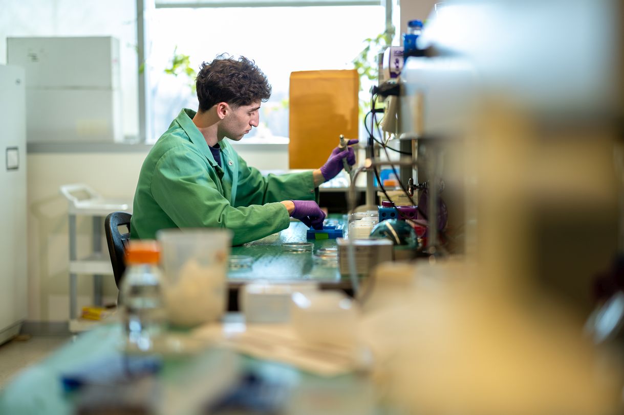 A young man with dark hair wearing a green lab coat and purple latex gloves uses a pipette at a lab bench.