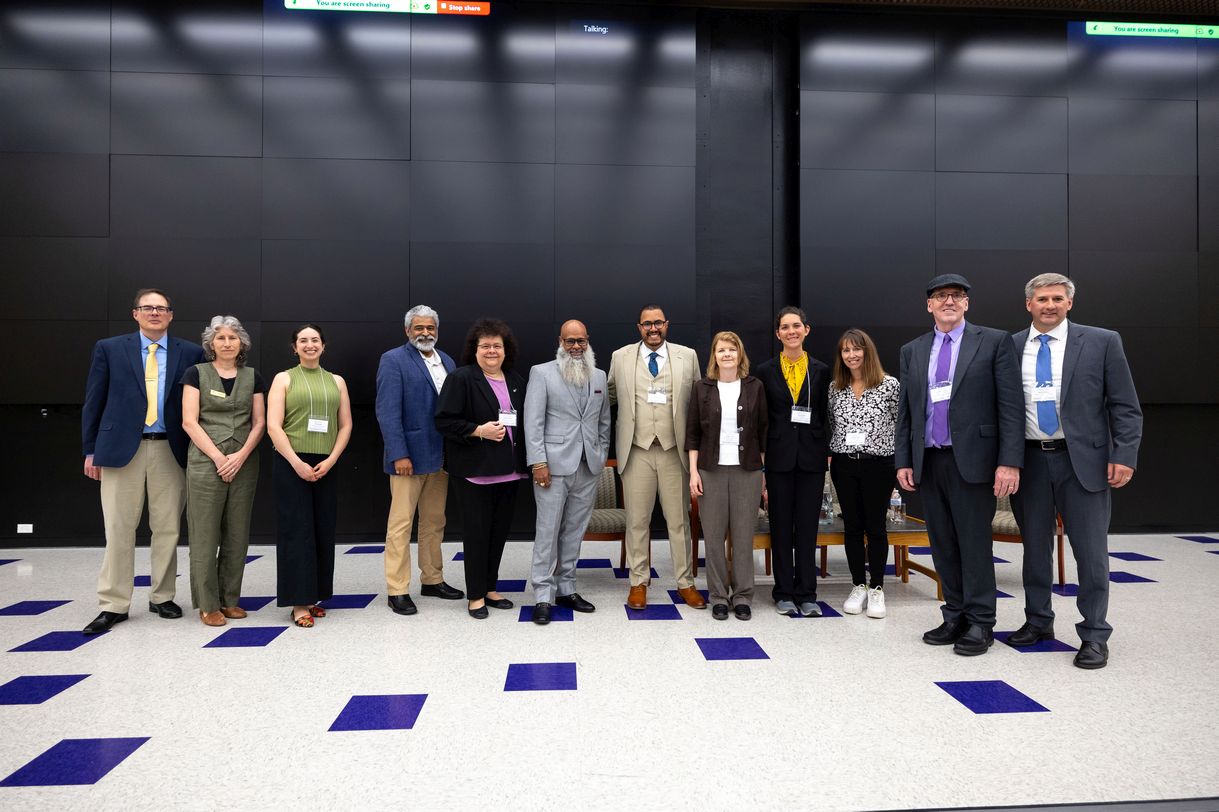 Twelve people stand in a line, posing for a group photo. All are smiling. They are standing against a wall covered in charcoal gray panels and the floor is covered in white and blue tiles. Those assembled represent faculty and staff from UAlbany’s School of Criminal Justice, Rockefeller College, and the Biology Department, with featured guests Steven Lopez and Eric Shapiro Renfroe.