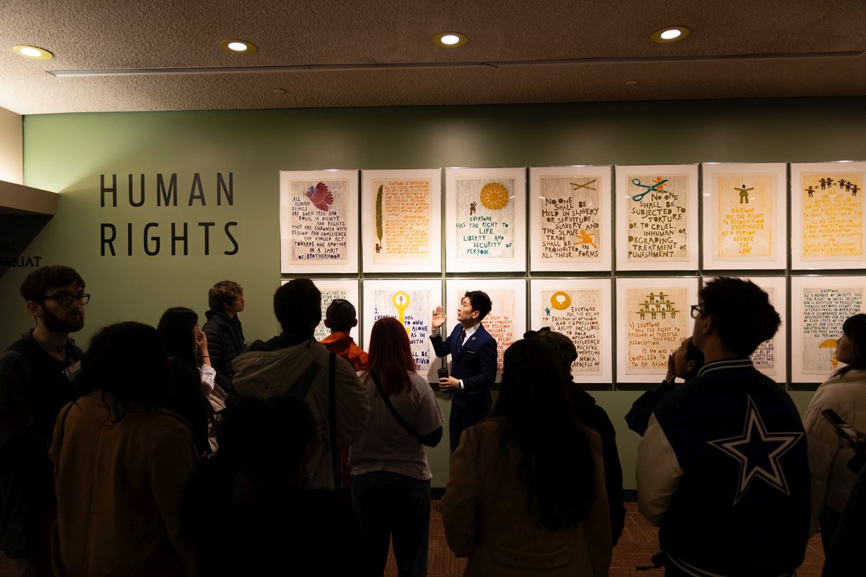 The image features a wall where 14 paintings are hung in two even rows of seven. To the left on the wall are the words ‘Human Rights’. A man wearing a blazer gestures to the paintings. A group of about a dozen students can be seen, in silhouette, in the foreground.  