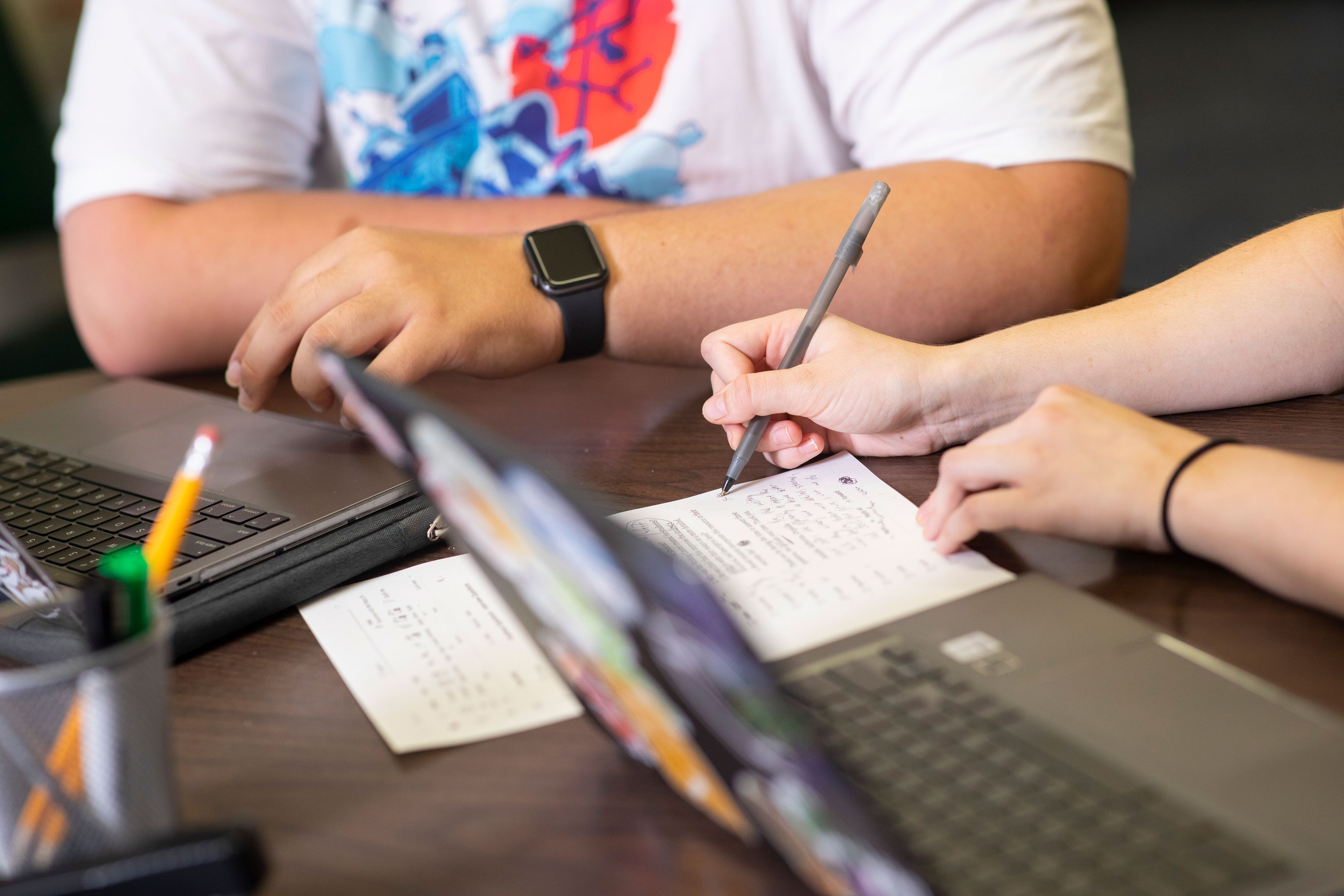 An image of two students sitting at a table with their computers open and one student is writing on a piece of paper.