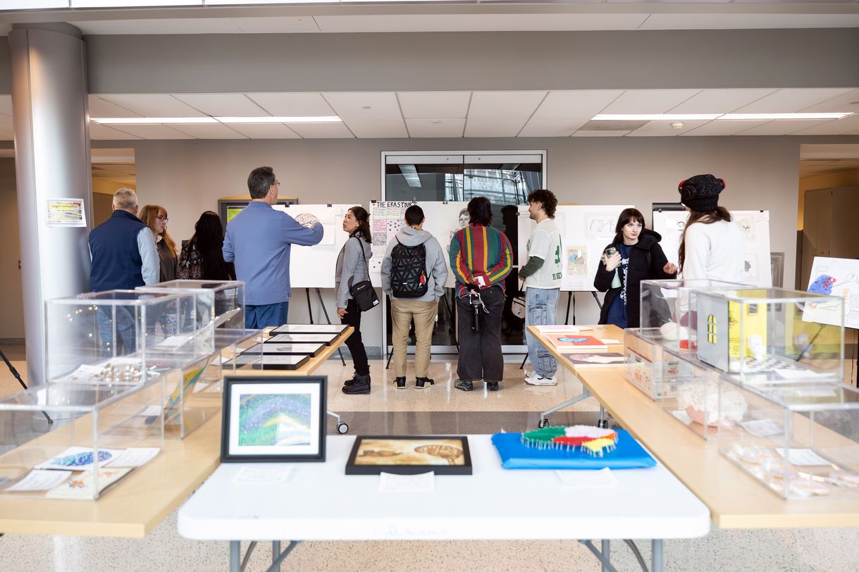 A group of people gather around easels displaying artworks. Other pieces are displayed on tables and in clear display cases in the foreground.