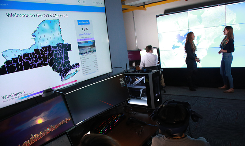 Faculty and students sit at computers and stand in front of a large screen with a map behind them at the xCITE (ExTREME Collaboration, Innovation and Technology) laboratory. (photo by Patrick Dodson)