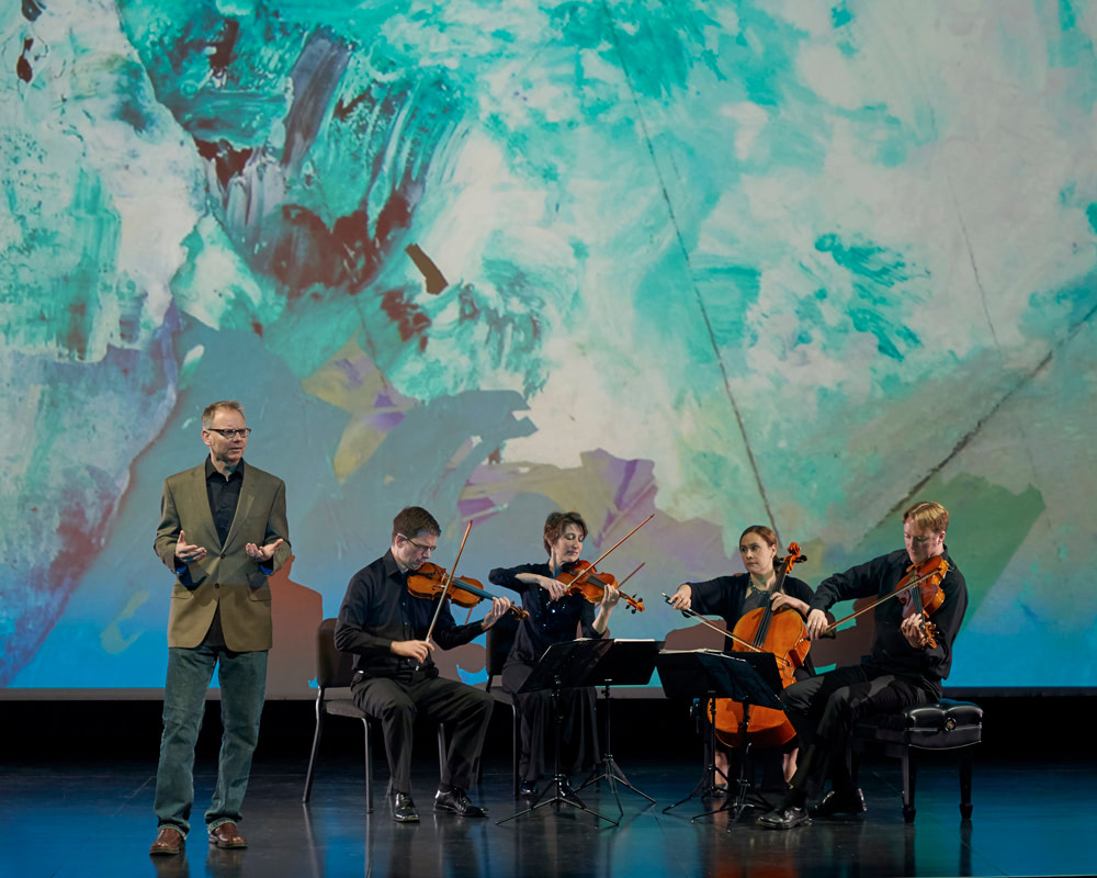 performer stands speaking by seated musicians playing instruments with image of clouds in background