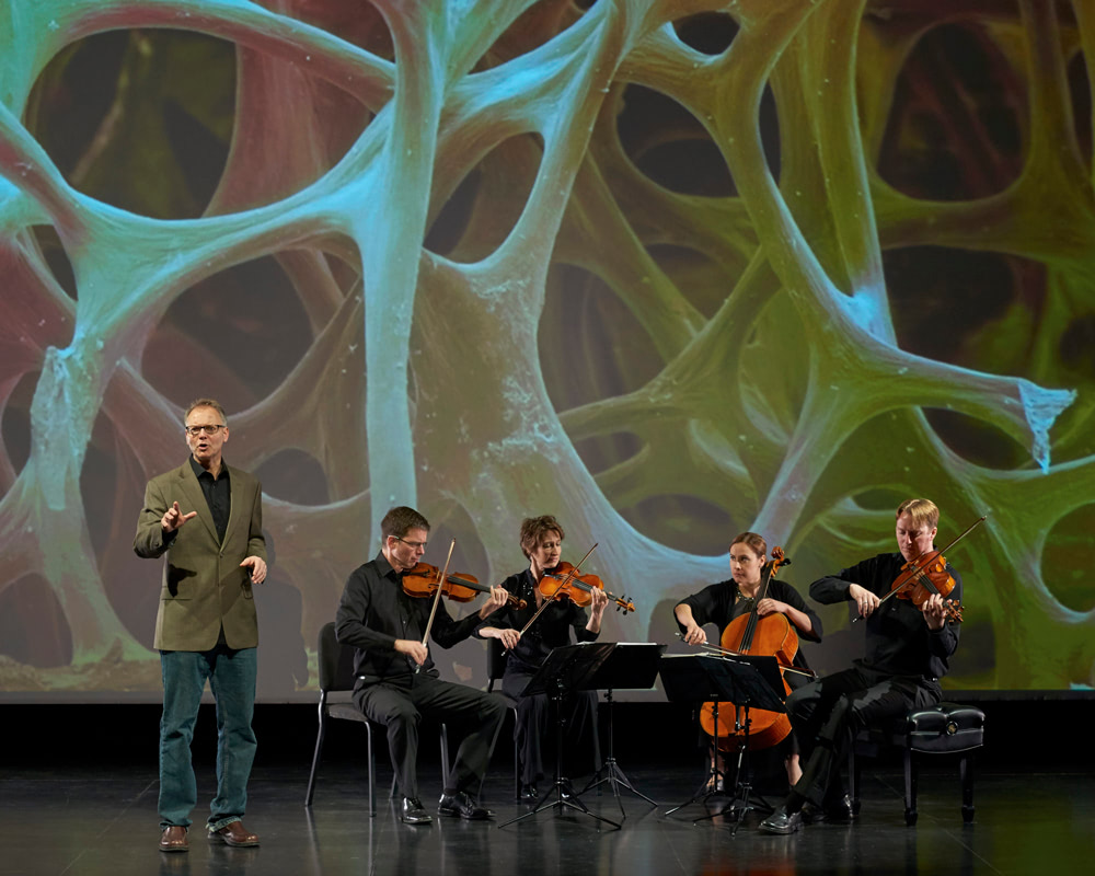 performer stands speaking by seated musicians playing instruments with image of DNA in background