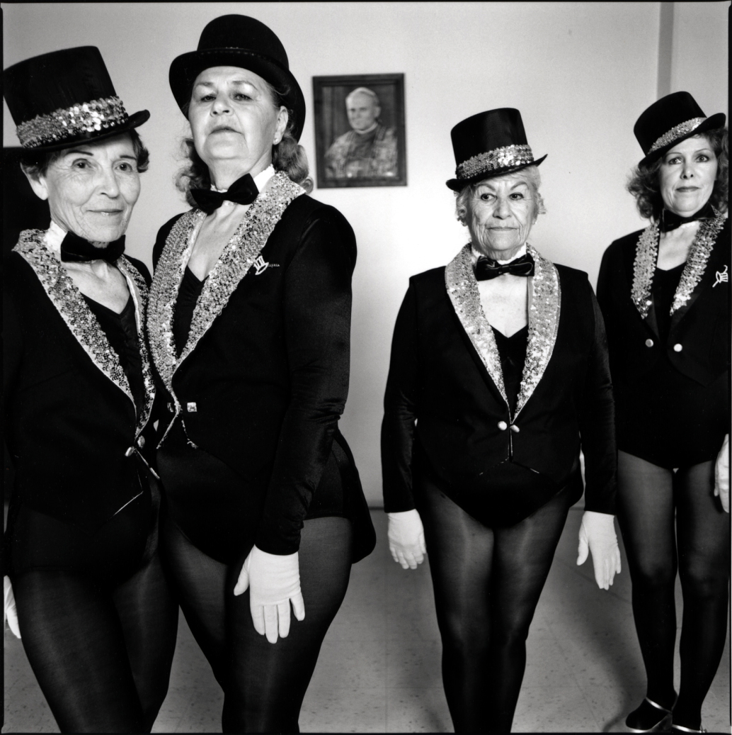 Four older women wearing costumes of sequined tuxedo jackets, tophats and tights stare at the camera