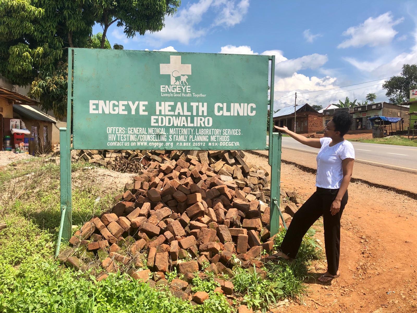 Elizabeth S. stands next to a green sign in Uganda that says "Engeye Health Clinic".