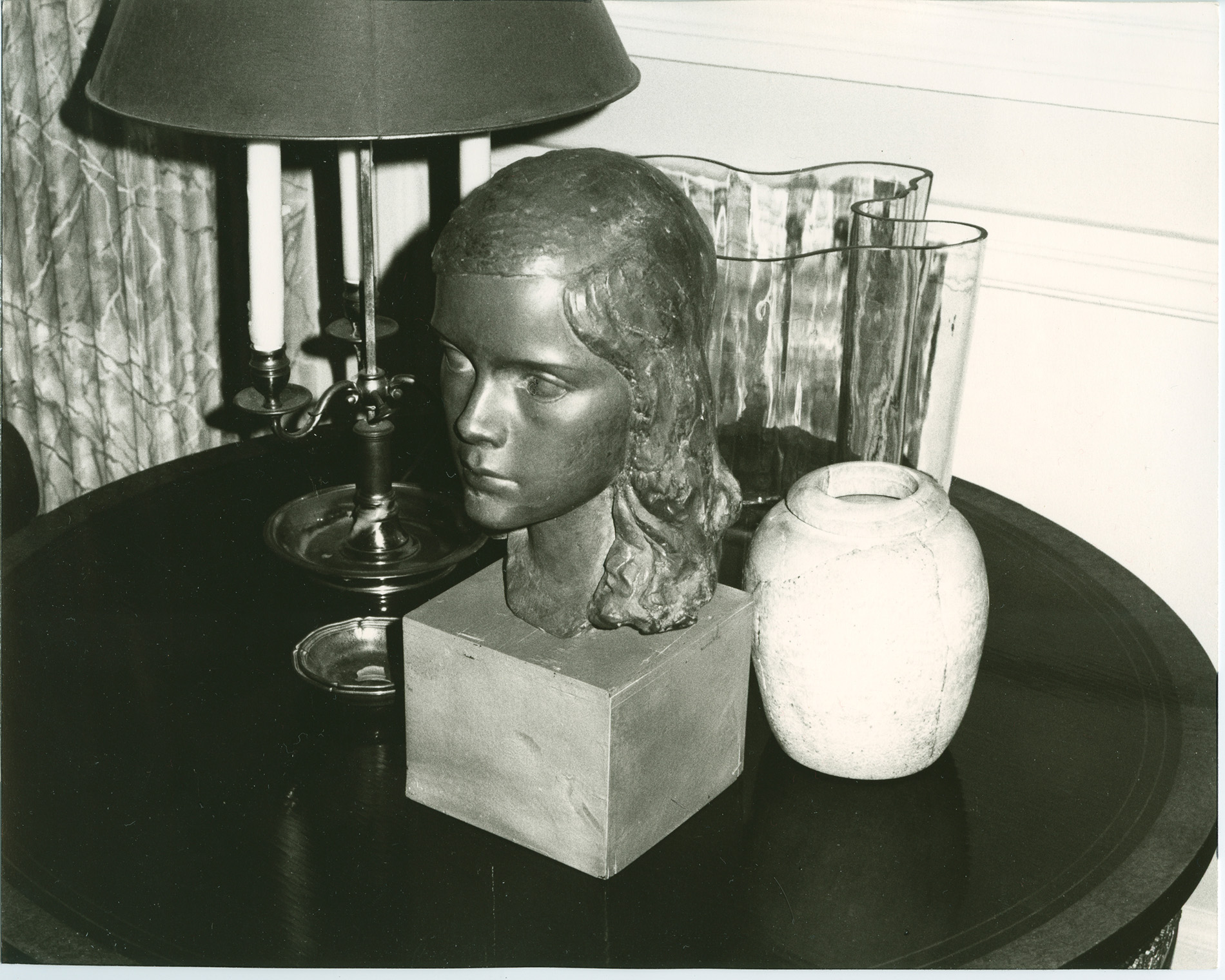 Black-and-white photo shows a table topped with a statue of a girl's head, vase and lamp. 