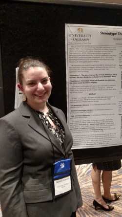 A woman in a gray blazer and a lanyard smiles with her hair pulled back in a bun. She is standing next to a research poster.