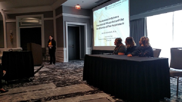 A woman in a black blazer stands behind a podium in a conference room to the left of a projection screen. To the right of the screen, three people sit at a table.