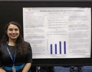 A woman with long curly brown hair stands and smiles to the left of a research poster. She is dressed in black and has a nametag around her neck.