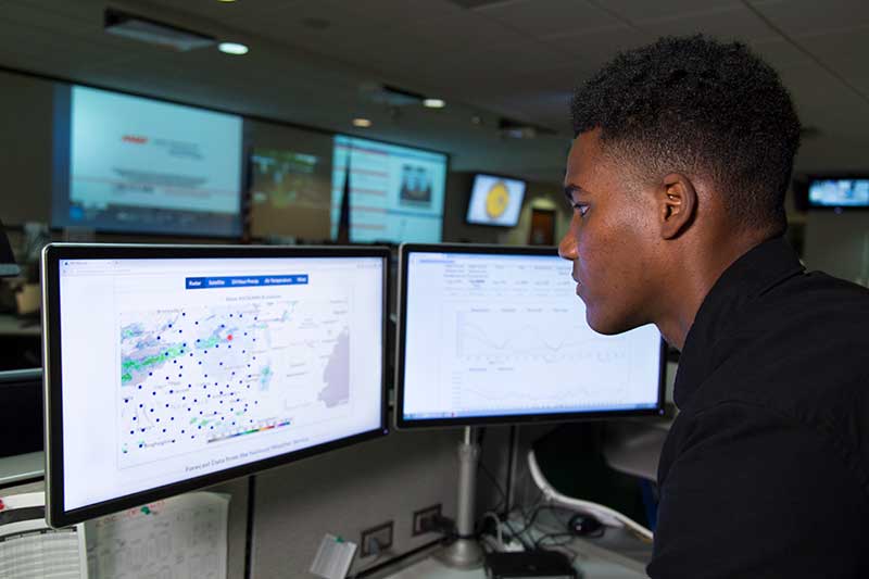 A man works on a computer with maps on the screen inside a command center.