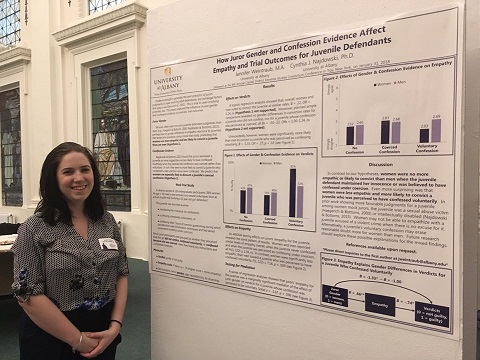 A brunette in a black and white blouse and a name tag smiles and stands next to a research poster mounted on a wall.