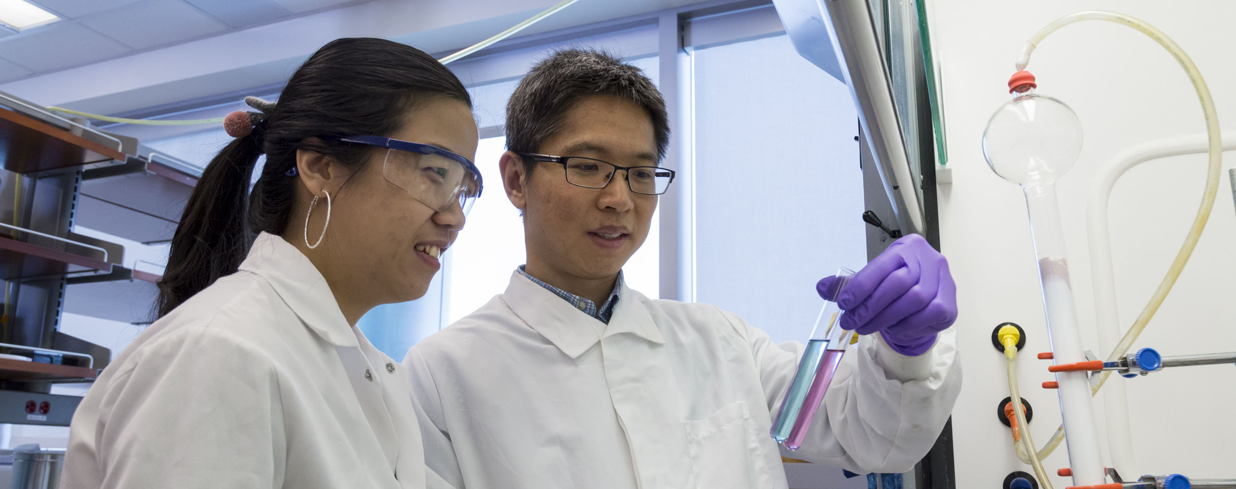Drs. Maksim Royzin and Jia Sheng, both of whom are wearing white lab coats and eye protection, look at a test tube with pink liquid and another with blue liquid in Dr, Sheng's hand.