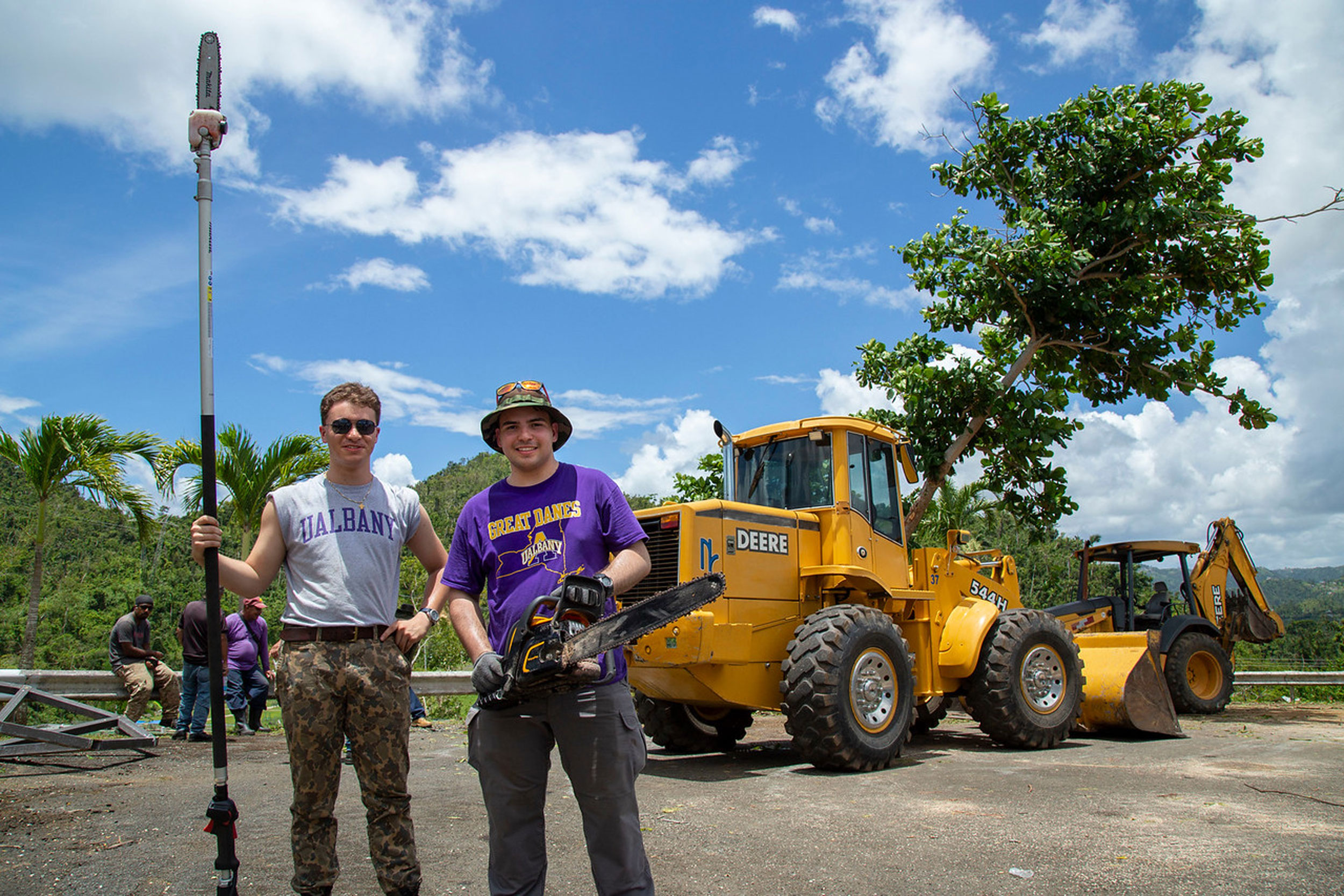 Two students pose with chain saws in front of heavy machinery while working in Puerto Rico after Hurricane Maria